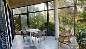 Een overdekt terras met een tafel en stoelen, grote ramen en uitzicht op groen.
