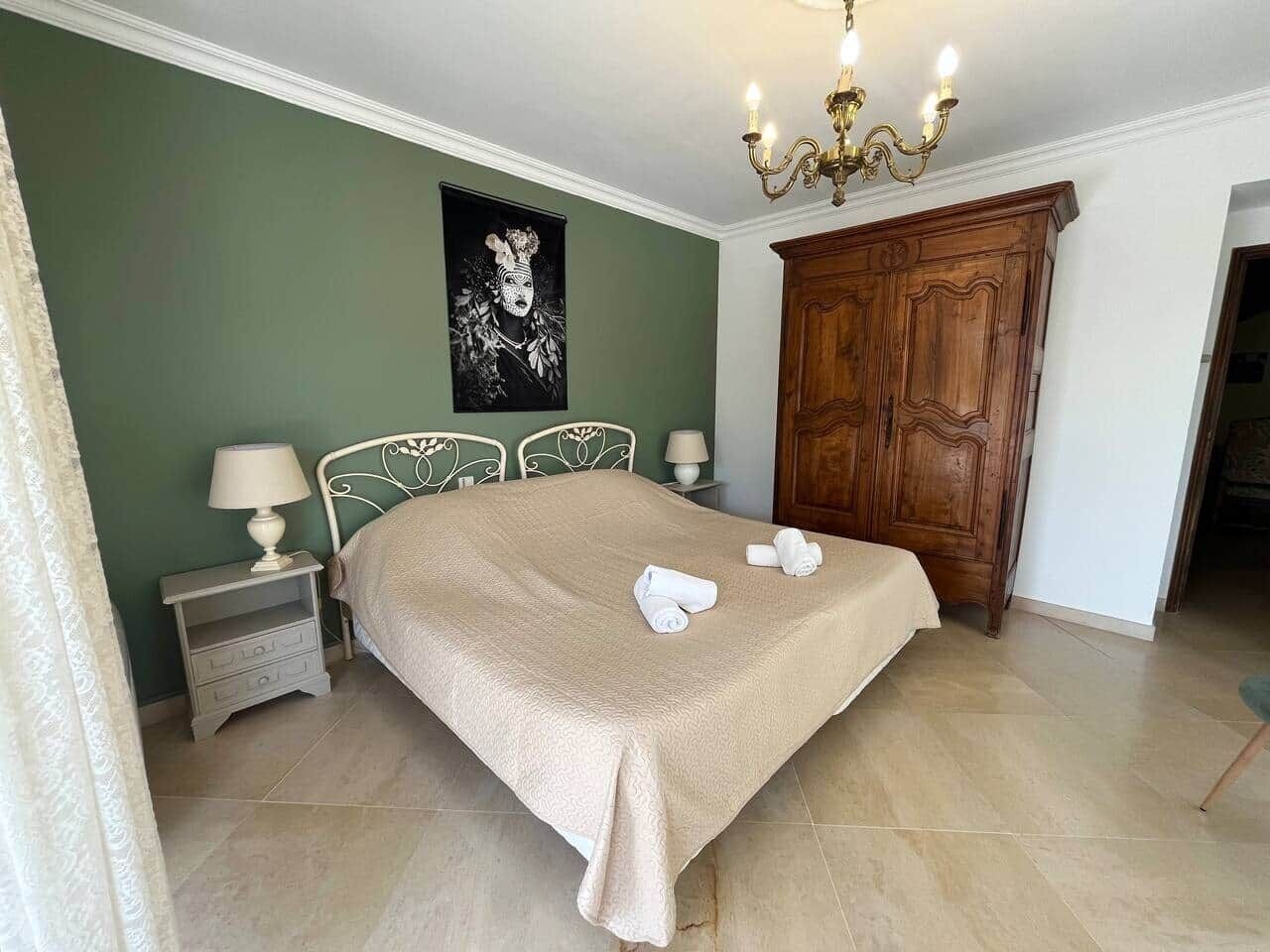 Ein ordentlich bezogenes Bett mit einer beigen Tagesdecke, zwei Nachttischlampen, einem hölzernen Kleiderschrank und einer Kandelaber.