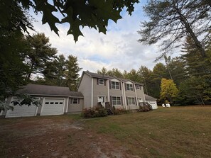 Une maison à deux étages avec un garage, entourée d’arbres et d’une cour gazonnée.
