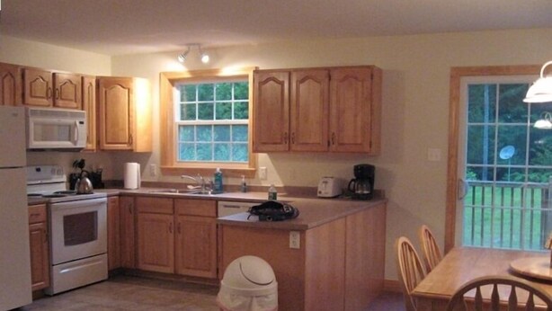 A kitchen with wooden cabinets, a microwave, a stove, a sink, and a dining area with chairs. - Sunday River Rental (Bethel)