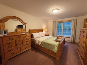 Ein Schlafzimmer mit einem hölzernen Bett, einer Kommode mit Schubladen, einem Spiegel, einem Fernseher auf der Kommode und einem Fenster mit Vorhängen.