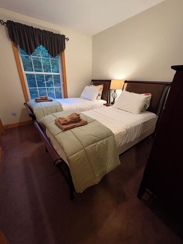 A hotel room with two beds, a window with curtains, a bedside table with a lamp, and a carpeted floor.