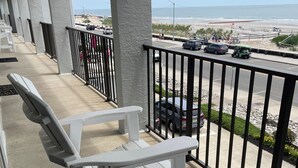A balcony with white chairs, a view of the beach, and a parking area with cars and a golf cart. - Direct Beachfront Condo With Great Ocean Views From Couch Or On The Porch (North Wildwood)