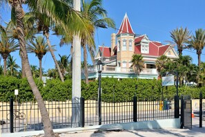 A large, historic building with a red roof and a tower, surrounded by palm trees and a fence.