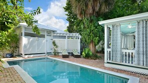 A pool area with a clear blue pool, a covered seating area with a ceiling fan, and lush greenery surrounding the space.