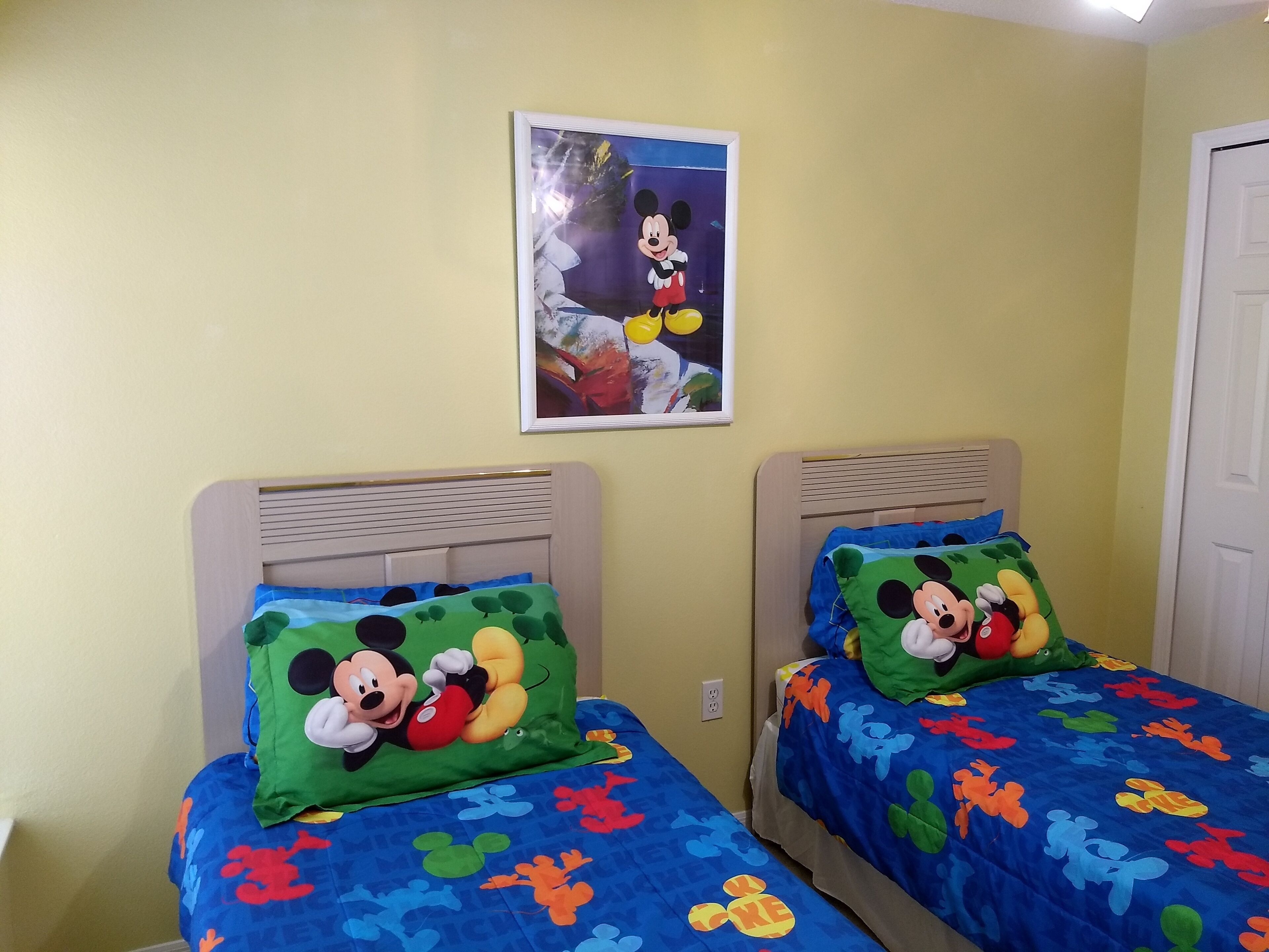 Deux lits jumeaux avec une literie aux motifs de Mickey Mouse, un tableau encadré représentant Mickey accroché au mur et un mur de couleur jaune unie.