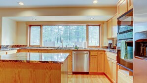 A modern kitchen with wooden cabinets, a large island, and a view of a forest through the windows. - 16 Yellow Pine - Gorgeous Golf Course, Covered Outdoor Living, Hot Tub, 3 King bedrooms, Bocce (Bend)