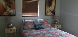 A bedroom with a patterned bedspread, two bedside lamps, a window with blinds, and two framed artworks on the wall.