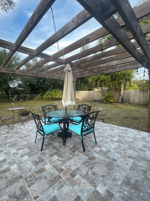 A covered patio with a round table and chairs, a pergola overhead, and a view of a grassy yard with a wooden fence.