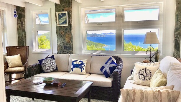 A cozy living room with a stone wall, a sofa with patterned cushions, a wooden coffee table, and a view of the ocean through large windows. - Mesmerizing view with pool 2 bedroom stone cottage in Coral Bay (St John)