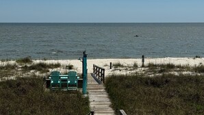 Ein hölzerner Steg führt zu Strandstühlen und einem Tisch, im Hintergrund das Meer.