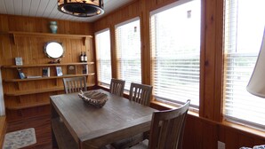 Ein Essbereich aus Holz mit Tisch, Stühlen und einer dekorativen Schale. Holzregale mit Büchern und Flaschen sind ebenfalls vorhanden, sowie ein Deckenventilator.