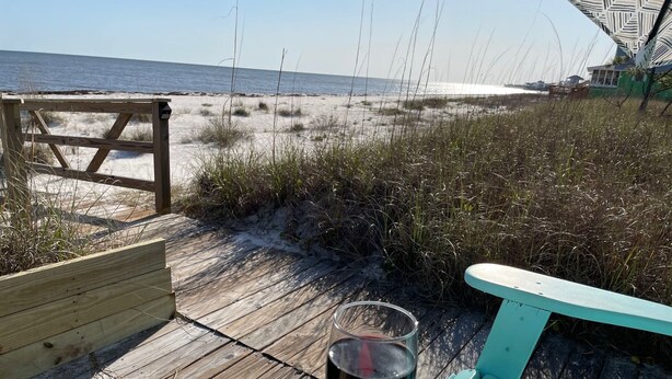 Eine Holzterrasse mit einem Glas Rotwein, einem türkisfarbenen Sessel und Blick auf Strand und Meer.
