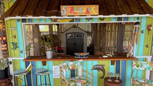 Eine farbenfrohe Tiki-Bar namens "Sure Shack" mit Holzfassade, einem Schild, das die Öffnungszeiten von 9:00 bis 20:59 Uhr anzeigt, und einer Speisekarte.