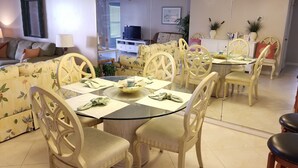 A dining area with a round glass table, white chairs, and a floral sofa in the background.