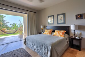 A bedroom with a large bed, two pillows, a nightstand with a lamp, and a view of a garden through a sliding door. - Amazing 2B apartment in Alegranza Luxury Condo, San José del Cabo. (Campo de Golf Fonatur)