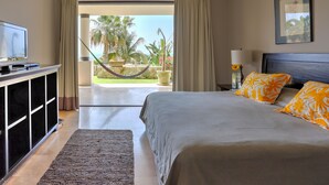Habitación de hotel con cama, televisión, escritorio y vista a palmeras y una hamaca afuera.