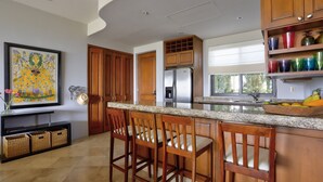 Una cocina moderna con armarios de madera, un refrigerador de acero inoxidable y una encimera de granito. Hay tres taburetes de bar de madera y una obra de arte enmarcada en la pared.