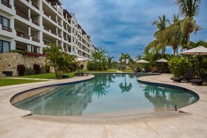 A swimming pool with a curved edge, surrounded by lounge chairs and umbrellas, with a multi-story building in the background. - Amazing 2B apartment in Alegranza Luxury Condo, San José del Cabo. (Campo de Golf Fonatur)