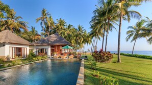 A swimming pool with lounge chairs and a thatched-roof building, surrounded by palm trees and a well-maintained lawn.