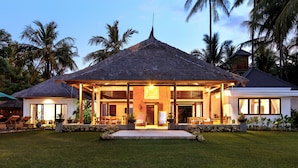 A beachfront villa with a thatched roof, a covered porch, and a well-maintained lawn.