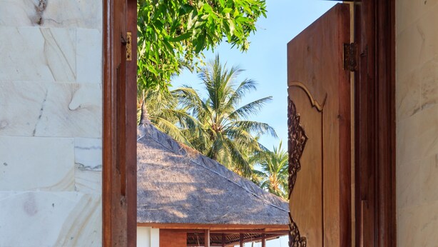 A wooden door leading to a covered outdoor area with a thatched roof, palm trees, and a stone pathway.