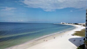 Une plage avec une eau d’un bleu limpide, un rivage sablonneux et quelques personnes qui se promènent. On aperçoit des bâtiments au loin.