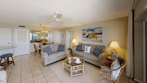 Un salon spacieux avec deux canapés, une table basse et des fauteuils en osier. On y trouve un ventilateur de plafond, un coin repas à l’arrière-plan et un tableau accroché au mur.