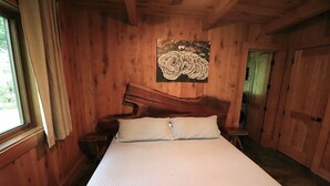 Un dormitorio de cabaña de madera con una cama, una mesita de noche, un cuadro en la pared y una ventana con cortinas.