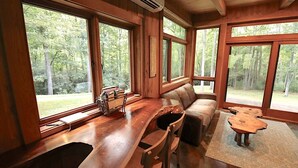 A cabin interior with a wooden dining table, a sofa, and large windows offering a forest view.