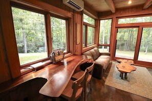Interior de una cabaña con una mesa de comedor de madera, un sofá y grandes ventanas que ofrecen vistas al bosque.