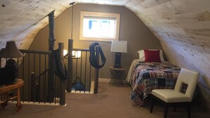 Une chambre mansardée chaleureuse, avec un plafond en pente, un lit recouvert d’une couverture à carreaux, une chaise, une petite table avec une lampe et un sac suspendu.