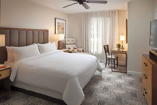 Una habitación de hotel con una cama grande, un escritorio, una silla, un ventilador de techo y un televisor.