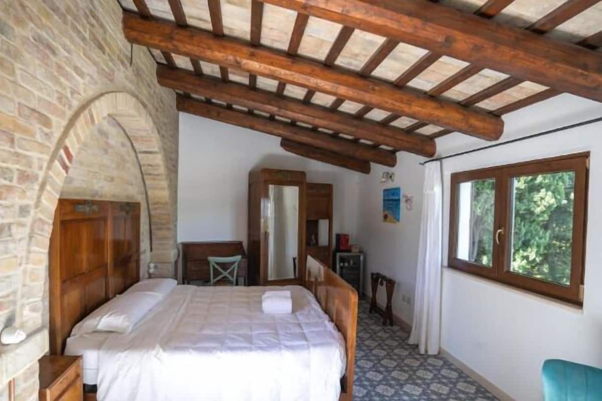 Ein Schlafzimmer mit Steinwand, Holzbalken, einem Bett, einem kleinen Tisch und einem Fenster mit Blick auf Grünfläche.