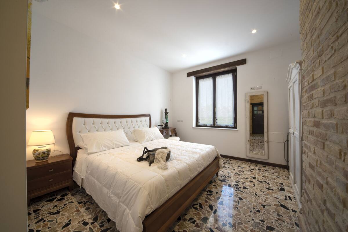 Ein Schlafzimmer mit Bett, Nachttisch, Lampe, Fenster und Spiegel.