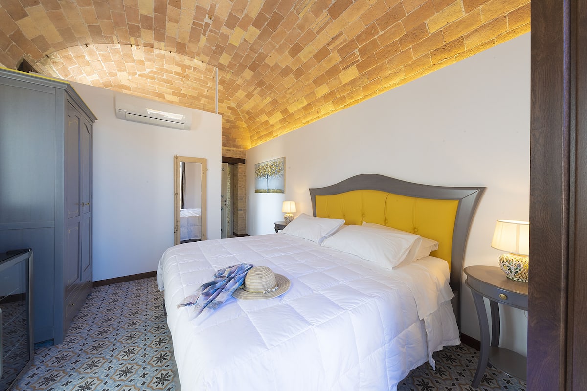 Ein Hotelzimmer mit Ziegeldecke, einem Bett mit weißer Bettwäsche, einem Nachttisch mit Lampe und einem gemusterten Boden.