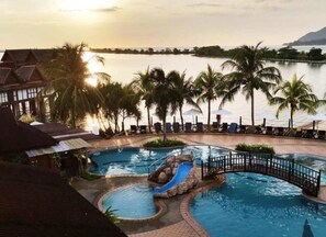 A swimming pool with a slide, palm trees, and a view of the water body at sunset. - De Water Chalet Langkawi Lagoon (Langkawi)