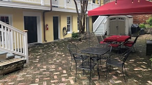 Un patio con suelo de ladrillo, mobiliario exterior con sillas de metal y mantel rojo, una sombrilla roja y un muro de ladrillo.