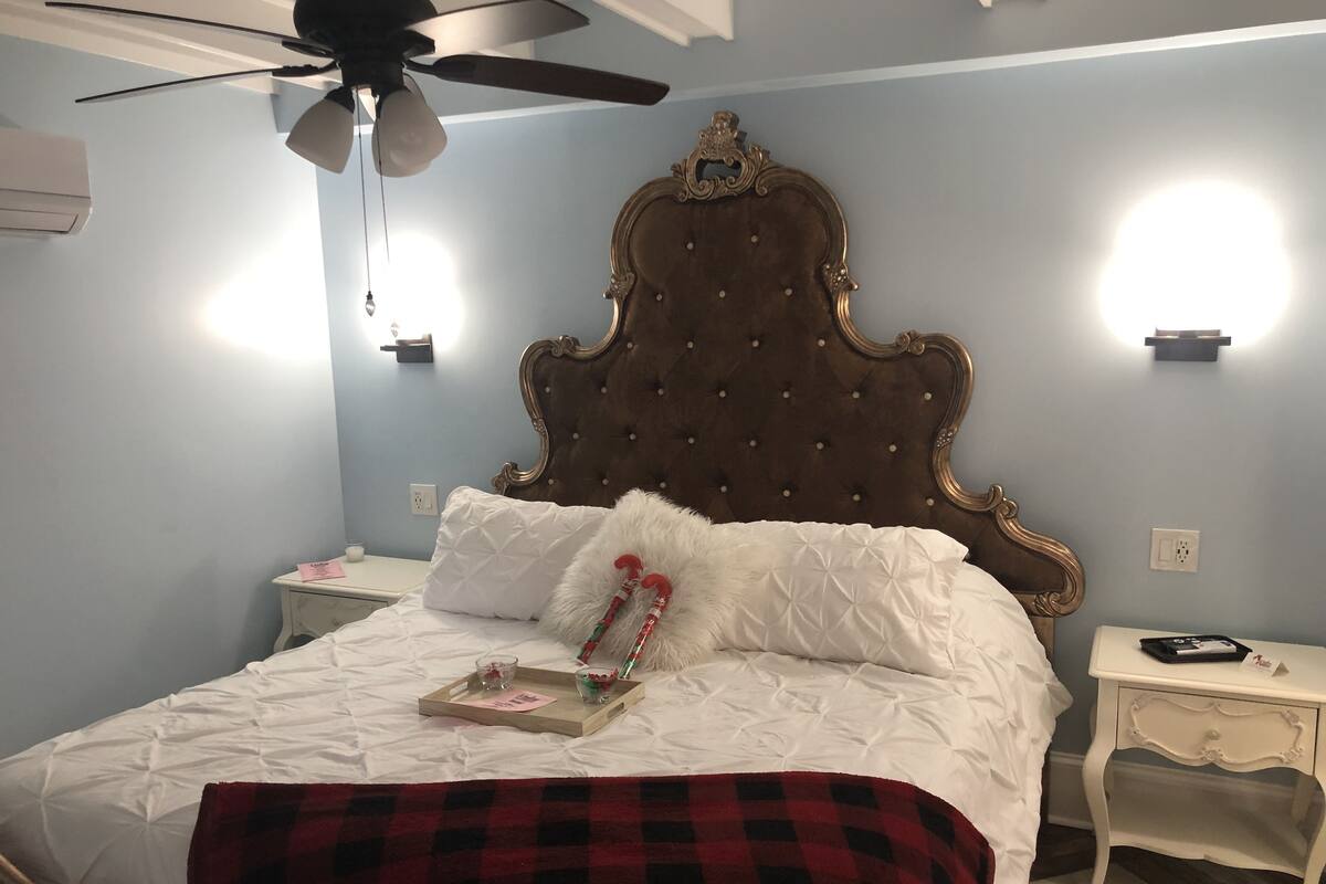 Une chambre à coucher avec une tête de lit capitonnée, un lit recouvert d’une literie blanche, une couverture à carreaux rouges et noirs, une table de chevet avec une lampe et un ventilateur de plafond.
