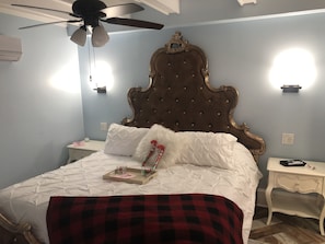 Un dormitorio con cabecera capitonada, cama con ropa blanca, una manta de cuadros rojos y negros, una mesita de noche con lámpara y un ventilador de techo.