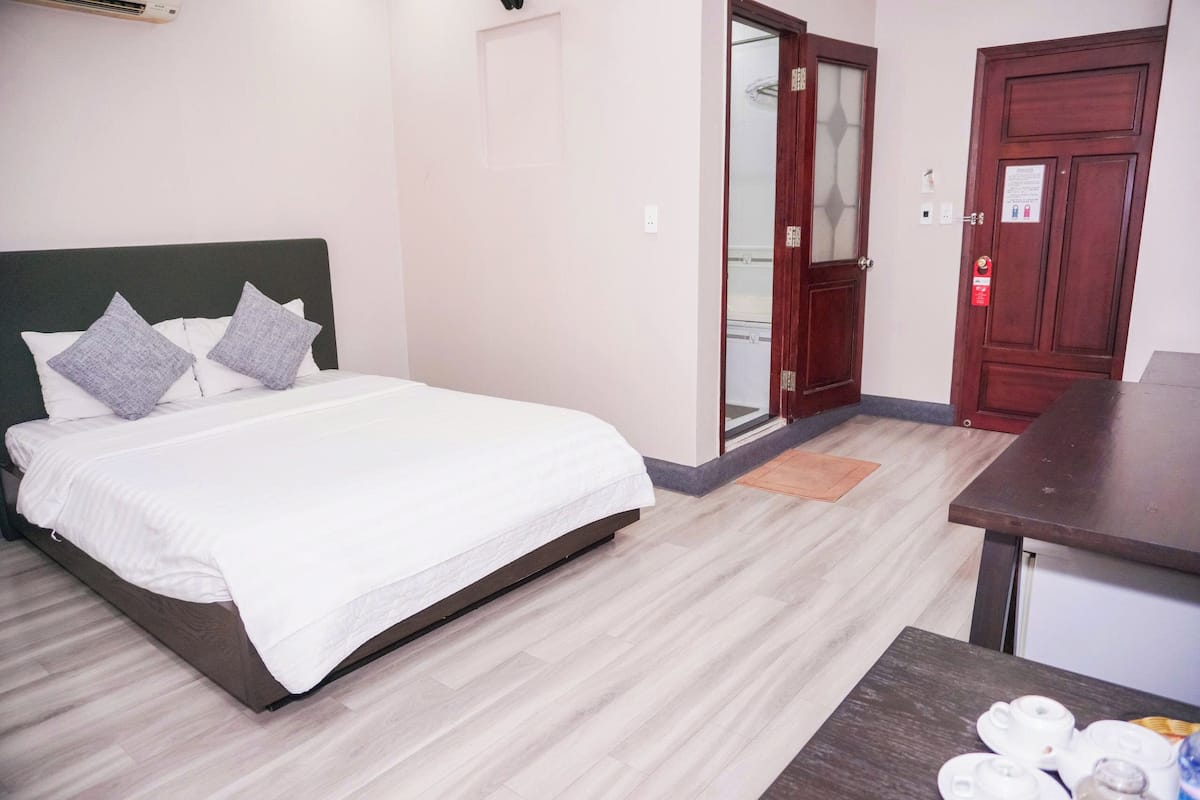 A hotel room with a bed, a wooden floor, a small table with cups, and a door with a glass panel.