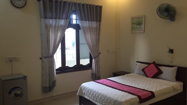A bedroom with a bed, a window with curtains, a wall clock, a small heater, and a wall-mounted fan. - Dang Tuan Hotel (Hue)