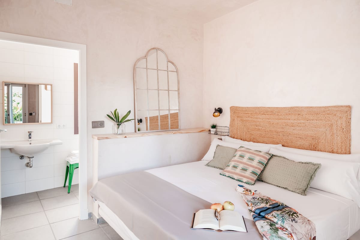 A bedroom with a bed, a bedside table, a mirror, a window, and a bathroom with a sink and toilet.