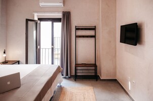 Una habitación de hotel con cama, escritorio con computadora portátil, televisor montado en la pared y balcón con cortinas.