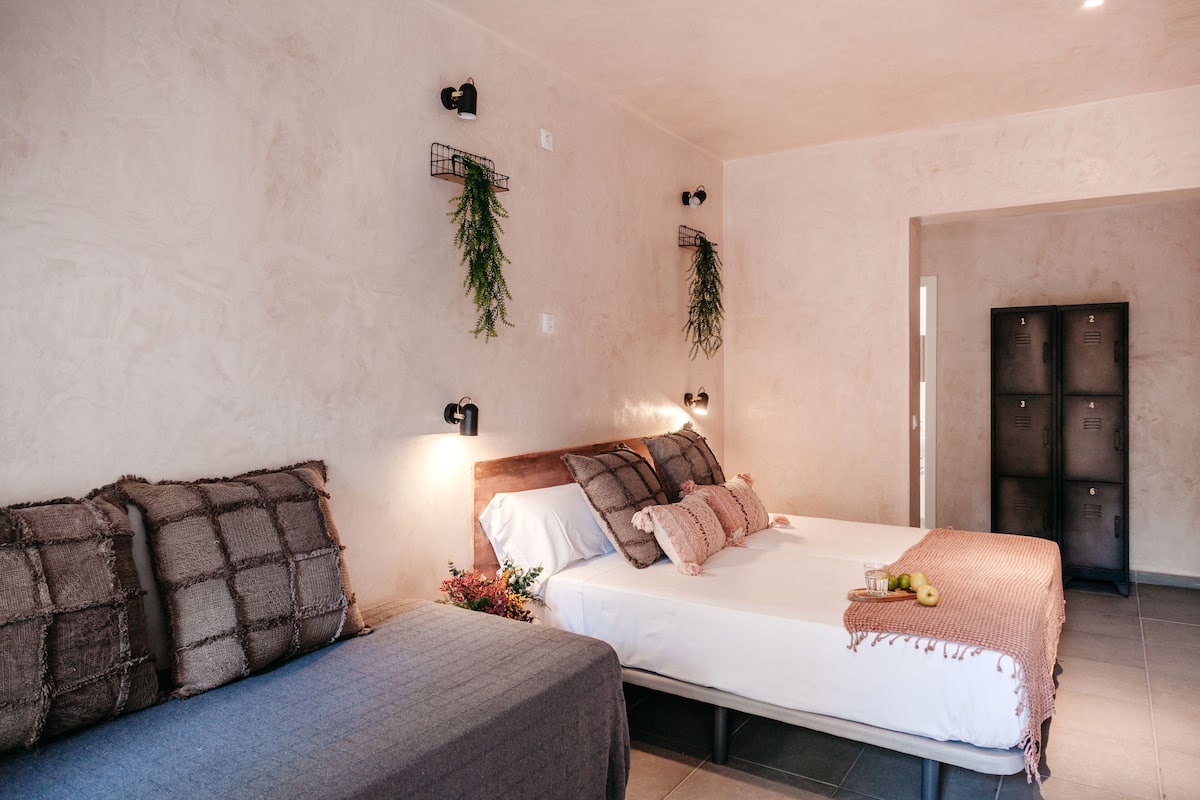 A bedroom with a bed, two bedside tables, a nightstand, a wall-mounted shelf with plants, and a door with numbered lockers.