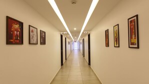 A long, well-lit hallway with framed artworks on the walls, leading to a bright end. - Hotel Luciyapalace (Thrissur)