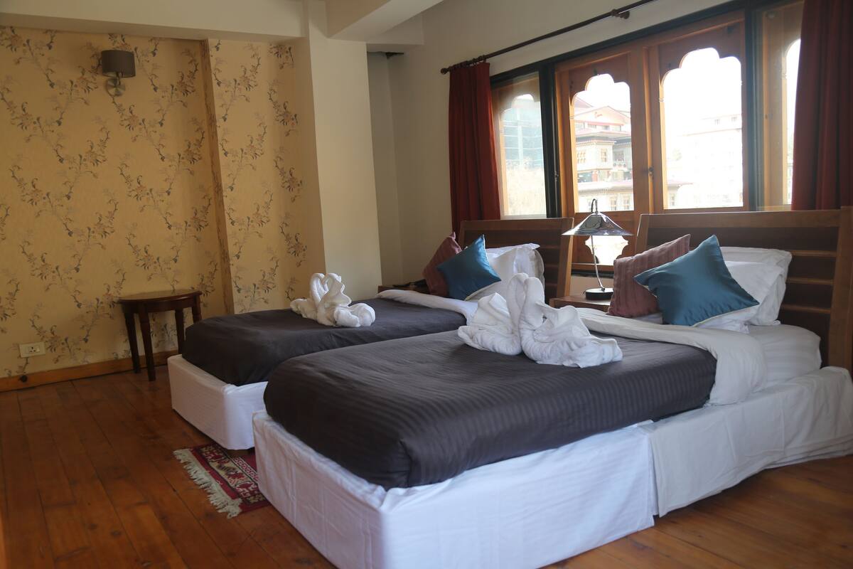 A hotel room with two beds, a wooden floor, a small table, and a window with curtains.