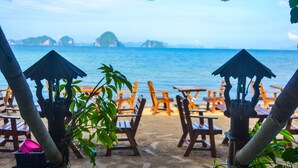 A beachside seating area with wooden tables and chairs, umbrellas, and a view of the sea.