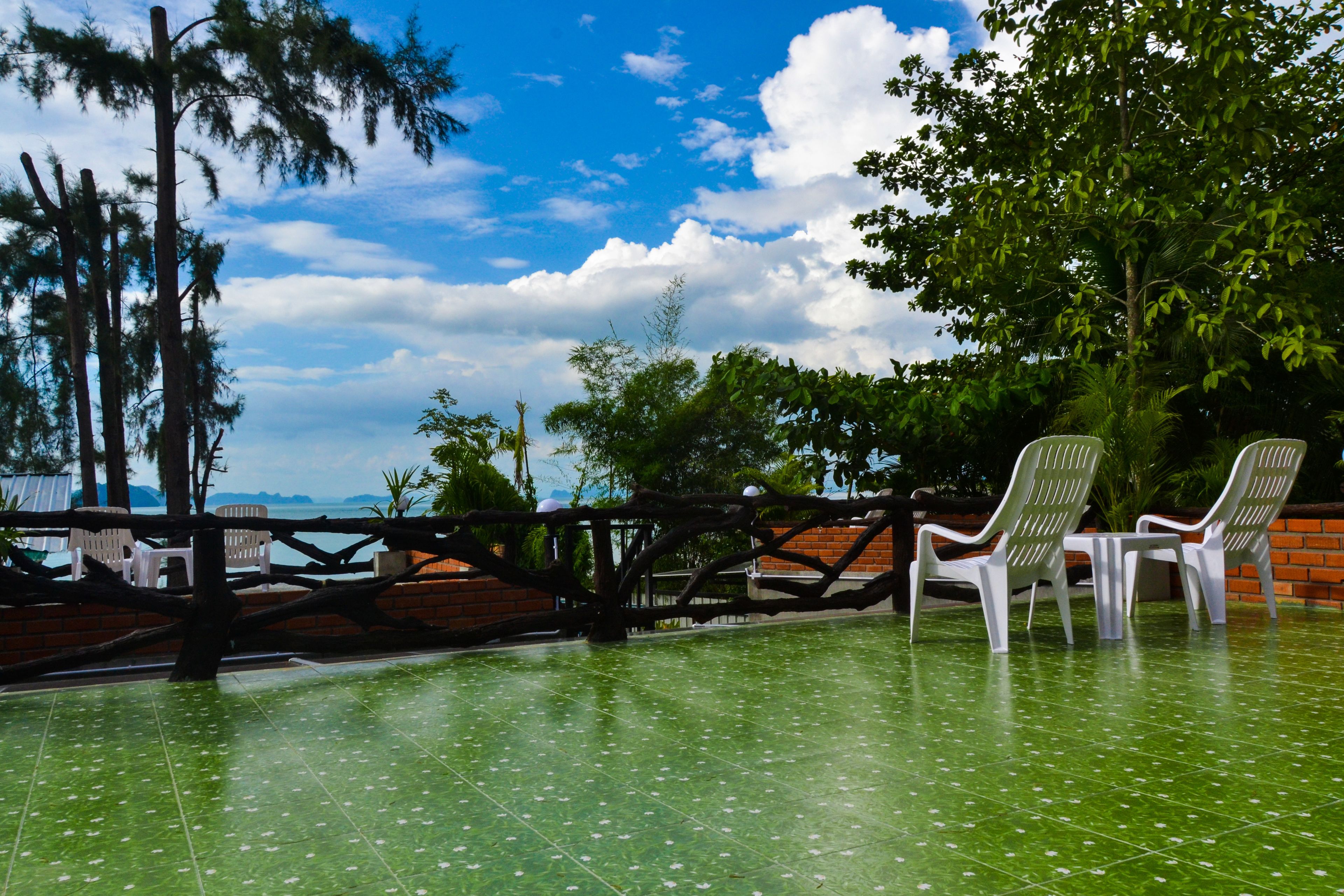 Une terrasse avec un sol carrelé de carreaux verts, deux chaises en plastique blanches, un mur en briques et une clôture en bois.