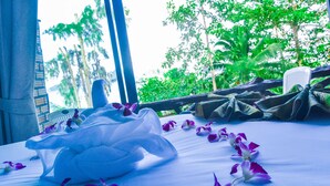 A bed with a white sheet and purple flowers, a window with a view of green trees, and a wooden chair.